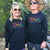 A female and Male standing in a garden wearing long sleeve black t shirts with a colourful graphic design for the Carnarvon Windfest