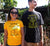 Female and Male in a garden, Female wearing yellow short sleeve t shirt with black design.  Male is wearing a black short sleeve t shirt with yellow design. Graphic of an outline of a tree with the text Trees are Free, Sow Seeds, Nurture by Nature.