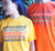 Female and Male wearing Short Sleeve T Shirts.  Female in golden yellow with graphic of stacked words in shades of black grey and red.  Male in orange with the graphics in brown, white and greenThe text reads Knowledge, repeated 3 times, and Powerful.