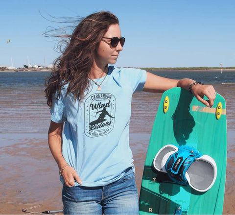 Female at a beach wearing a light blue fitted short sleeve t-shirt.  Design is in black.  The graphics are of a silhouette of a kite surfer with the text Wind Riders, Carnarvon Western Australia.