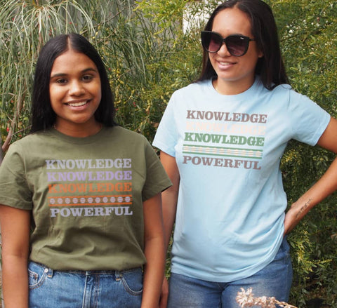 Two Females in a garden wearing Fitted Short Sleeve T Shirts.  One in a khaki t-shirt with graphics is stacked words in shades of purples and orange. One in a light blue t-shirt with graphics is stacked words in shades of brown and grenn.  The text reads Knowledge, repeated 3 times, and Powerful.