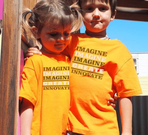 Boy and Girl outside with arms around eachother wearing orange Short Sleeve T Shirts.  Graphic is stacked words in shades of brown and white.  The text reads Imagine, repeated 3 times, create, innovate.