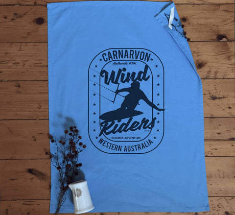 Light blue tea towel flay lay in a kitchen.  Design is in black.  The graphics are of a silhouette of a kite surfer with the text Wind Riders, Carnarvon Western Australia,