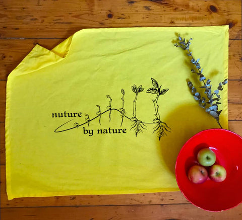 Flat lay Yellow Tea Towel in a kitchen. The design is in black. The graphic is an outline image of a seed growing in stages. The text is nurture by nature.
