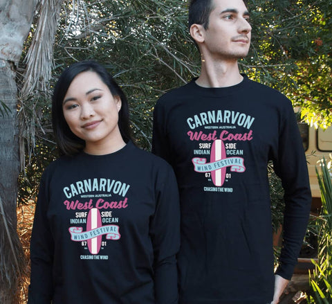 Male and female in a garden wearing black Long Sleeve T Shirts.  Design in blue and pink.  Surfboard with banner saying Wind Festival.  Text reads Carnarvon Western Australia.  West Coast. Surf Side. Indian Ocean. 6701. Chasing the wind.