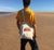 Man on a beach using a long handle calico bag with graphic design of a silhouette of a kelpie dog with the text Best Mate on the Side of Adventure