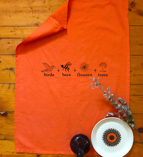 Flat lay Orange Tea Towel in a kitchen. The design is in black. The graphics are 4 outline images with words underneath and a plus sign in between. The images are of a bird, bee, flower and tree.