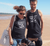 Female and Male on a beach with a dog wearing black singlet tank t shirts.  Design in white.  Graphic is a silhouette of a kite surfer with the text Carnarvon Western Australia.  It's all About the Summer Wind.