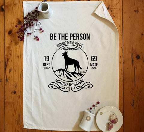 Flat Lay Natural colour Tea Towel with design. The graphic is one colour.  There is a circle outline with a silhouette of a dog standing on a rocky outcrop.  Around the circle the text says - Be the person your dog thinks you are, Nurture by Nature.  On either side of the circle there is text that is split - it says 19-69, Best Mate, Wild Life