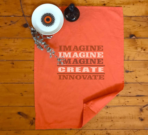 Orange Tea Towel.  Graphic is stacked words in brown and shades of beige.  The text reads Imagine, repeated 3 times, create, innovate.