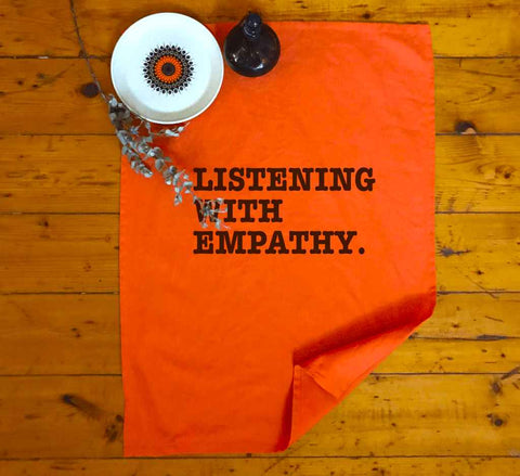 Orange Tea Towel flat lay in a kitchen. Graphic is stacked words in black. The text reads Listening with Empathy.