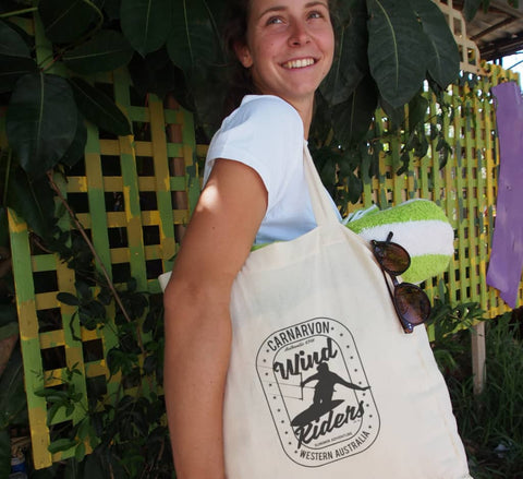 Female using a calico shopping bag at a beach wearing a white t shirt fitted short sleeve t-shirt.  Design in black is on the calico.  The graphics are of a silhouette of a kite surfer with the text Wind Riders, Carnarvon Western Australia.