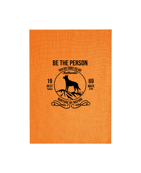 Orange Tea Towel with design. The graphic is one colour.  There is a circle outline with a silhouette of a dog standing on a rocky outcrop.  Around the circle the text says - Be the person your dog thinks you are, Nurture by Nature.  On either side of the circle there is text that is split - it says 19-69, Best Mate, Wild Life