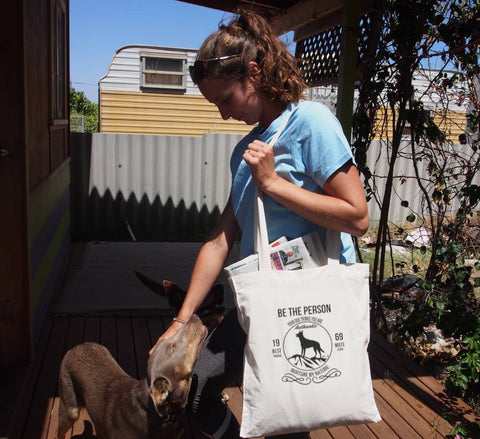 Female patting dog, outside a house, using a Long handle calico bag with the design. The graphic is one colour.  There is a circle outline with a silhouette of a dog standing on a rocky outcrop.  Around the circle the text says - Be the person your dog thinks you are, Nurture by Nature.  On either side of the circle there is text that is split - it says 19-69, Best Mate, Wild Life