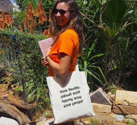 Female in a garden using a Long Handle Calico Bag, natural colour.  Graphic in Black.  Think Flexibly repeated in 4 lines written in all directions..