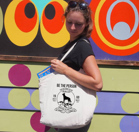 Female outside using a short handle, shopping style calico bag with the design. The graphic is one colour.  There is a circle outline with a silhouette of a dog standing on a rocky outcrop.  Around the circle the text says - Be the person your dog thinks you are, Nurture by Nature.  On either side of the circle there is text that is split - it says 19-69, Best Mate, Wild Life