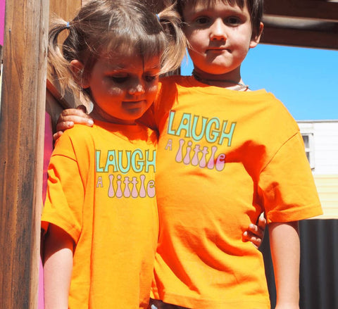 Boy and a girl outside wearing orange Short Sleeve T Shirts. Graphic is stacked words in pale green and pale purple with black outline. The text reads Laugh a Little.